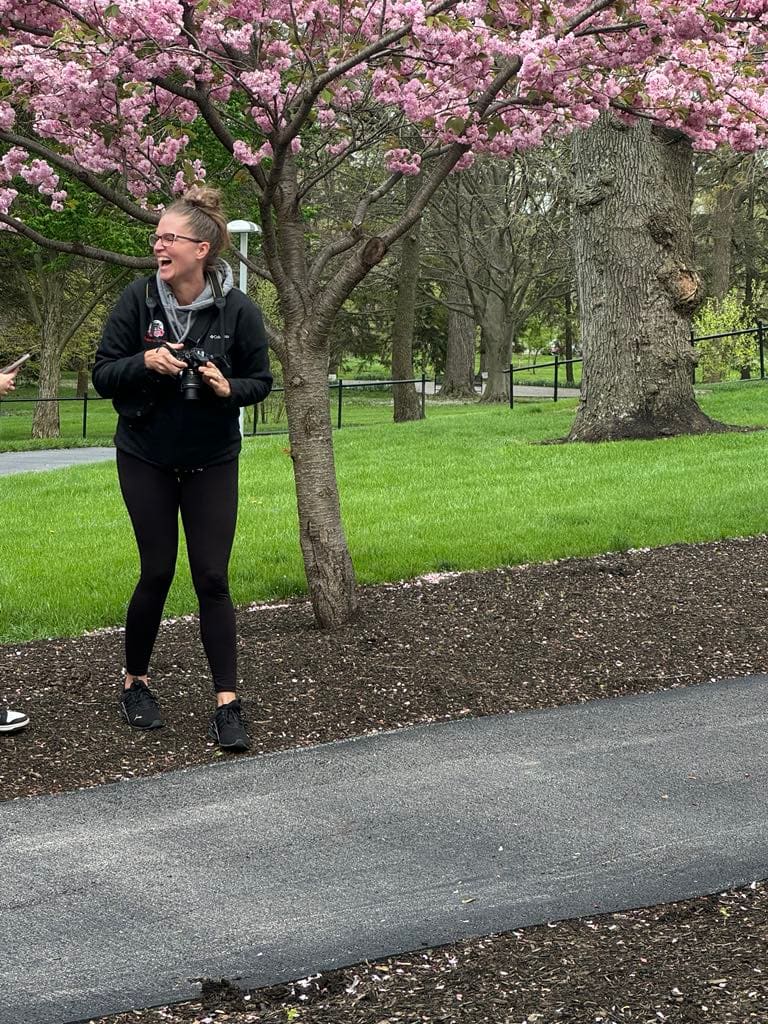 Photographer Betsy laughing while holding DSLR camera under pink cherry blossom tree in park